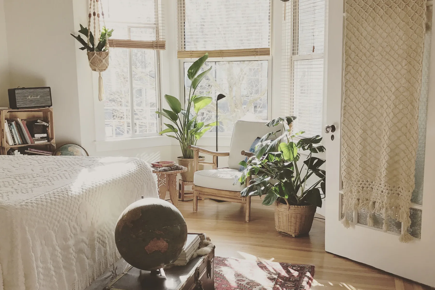 Dormitório com roupa de cama clara, poltrona de madeira, plantas e luz natural.